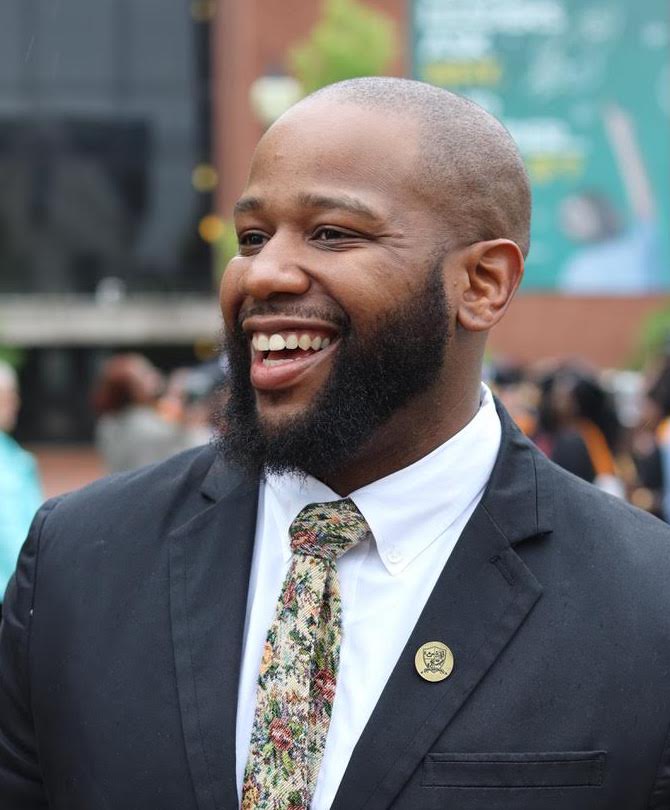 Jamaal Gosa, a smiling Black man with a shaved head and full beard wearing a black suit and floral tie
