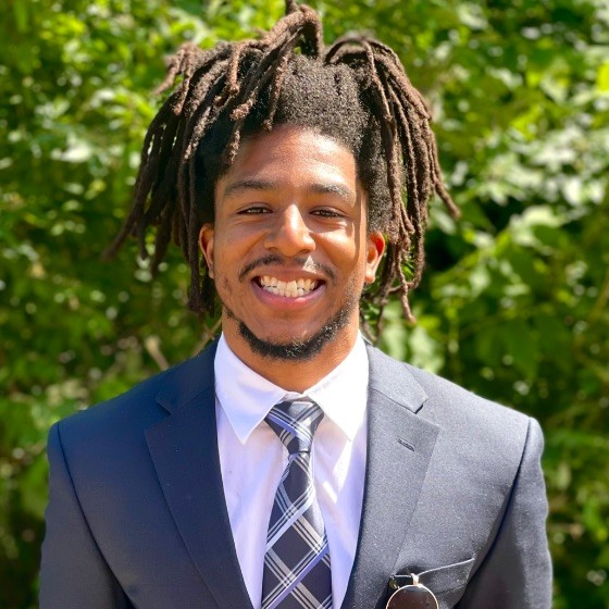 Rodney Gaskins-Acie, a smiling African American man wearing a black suit with plaid tie and hair in locs in a ponytail