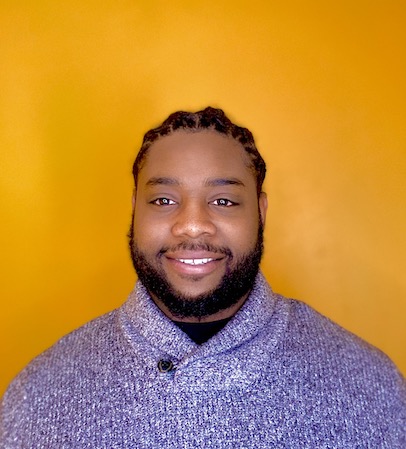 Spencer Scott, a smiling Black man with hair pulled back in braids and a full beard wearing a blue knit sweater.